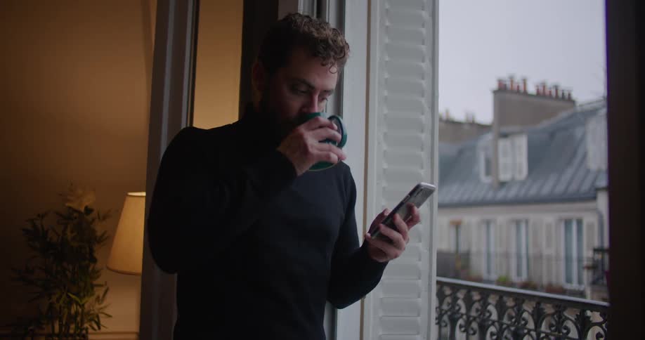 A man is standing inside near a balcony making a call on his cell phone as he drinks from a cup