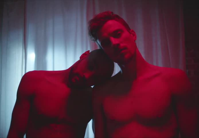 Men standing barechested in room lit by red lighting lean their heads against each other