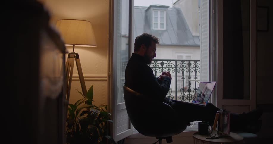 A man sits in front of his balcony at home and video chats on his laptop