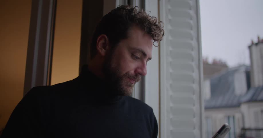 A man drinks coffee while scrolling through his phone on his balcony