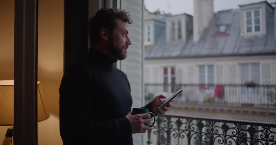Man stands on balcony drinking coffee and texting on phone