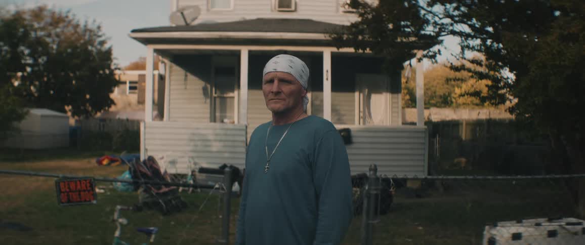 Man wearing bandana stand in front of fence gate with house in background