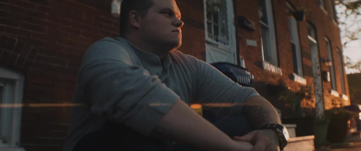 Man sits on curb near house as sun glares across street from the horizon