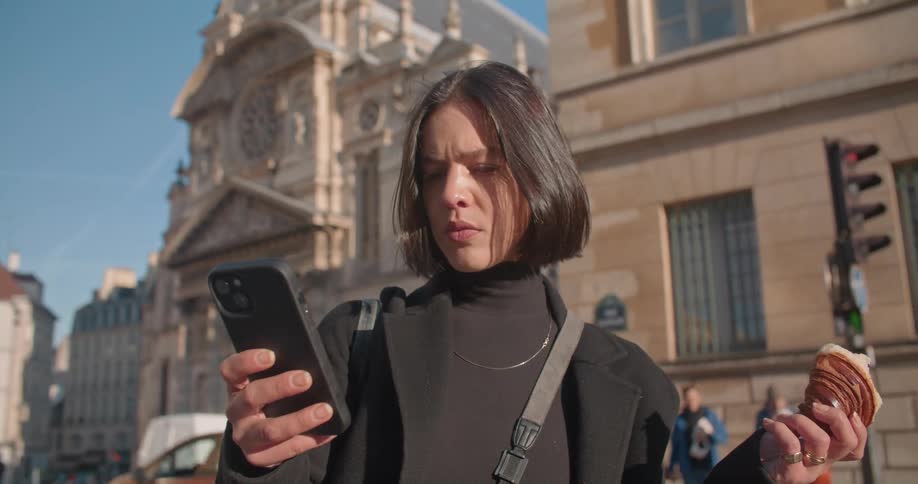 Woman with short straight hair black turtleneck sweater and blazer uses smartphone eating croissant and looking around in the city center