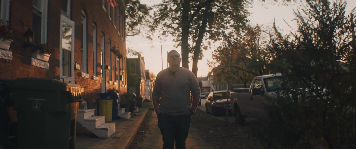 Man with grey sweater and jeans walks down the street next to apartment buildings at sunset