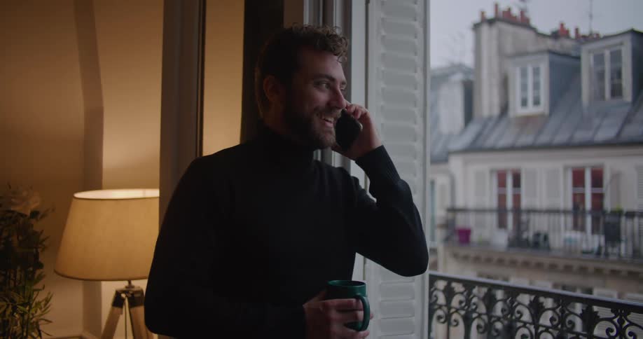 Young man wearing a black sweater holding a mug talking on the phone near the apartment balcony