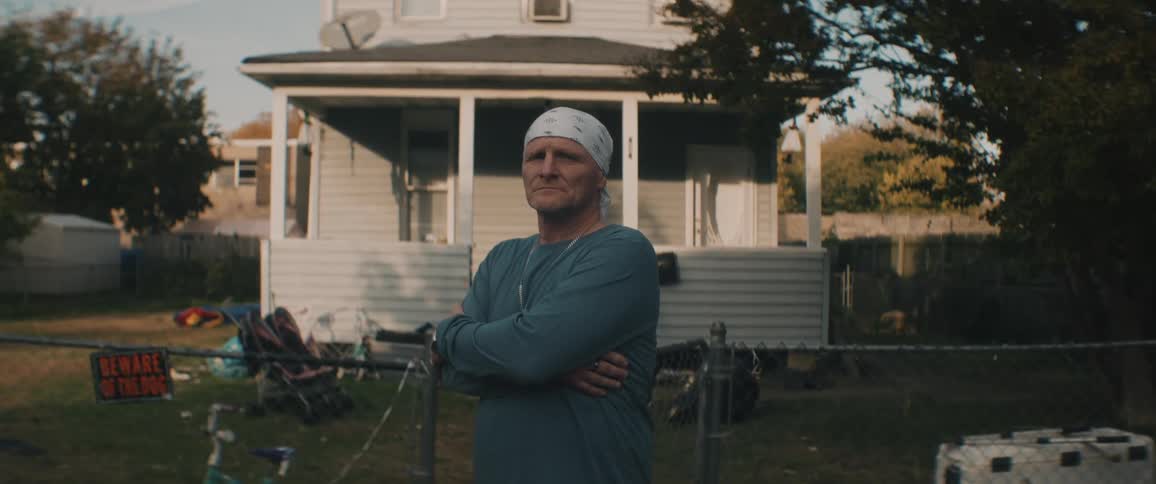 Man with a bandana on his head stands with his arms crossed near open gate in front of house