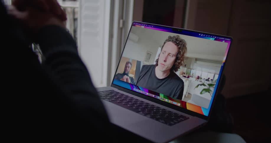 A man chats with a friend via video call on his laptop while sitting next to his window