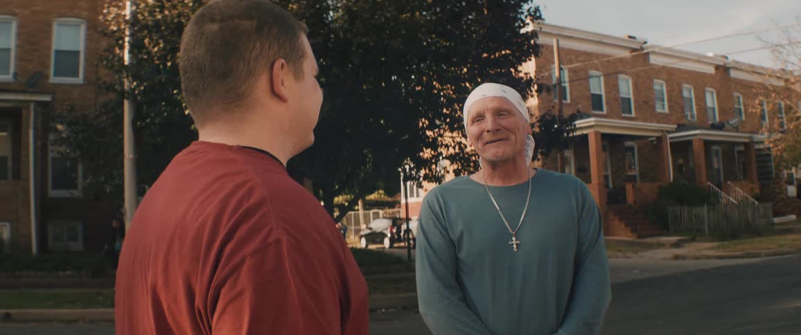 A man wearing a white bandana on his head is standing on a neighborhood street talking to a young man and laughing