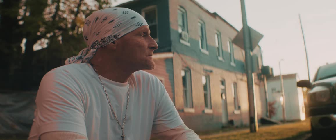 Man wearing bandana sits on neighborhood curb near parked vehicle