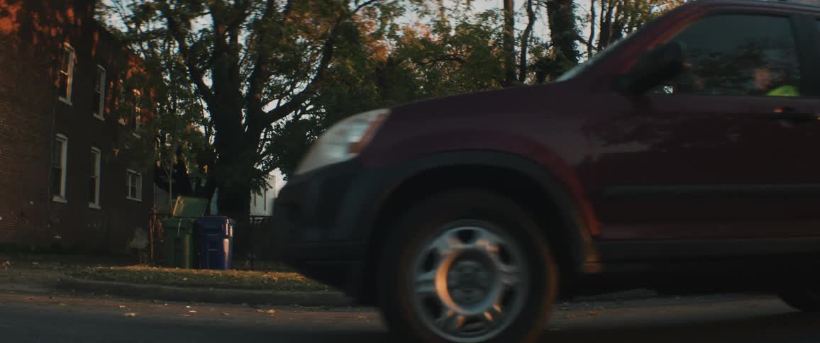 A man sits on a curb in a neighborhood as a vehicle drives past him