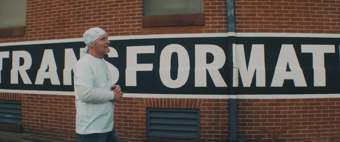 A man walks down a sidewalk next to a large sign and smiles and talks