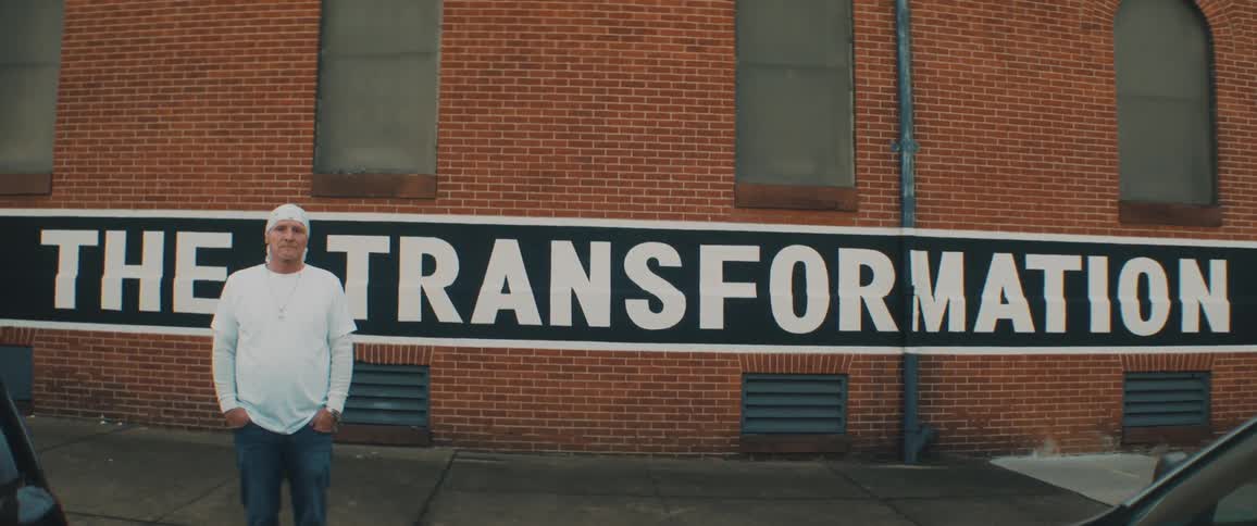 A man stands with his hands in his pockets on a sidewalk in front of a building with a large sign