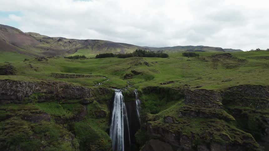 People walk on the paths beneath green and rocky cliffs as a waterfall flows down