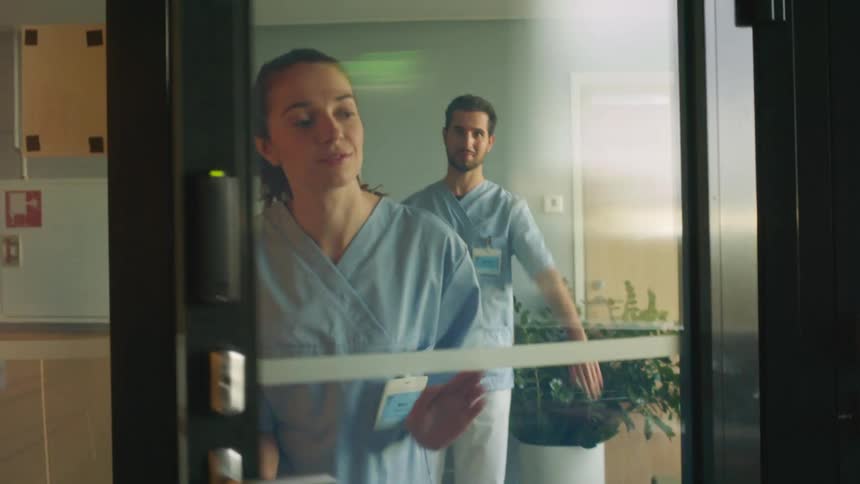 A male nurse waves to a female nurse as she is walking out a door in a medical center