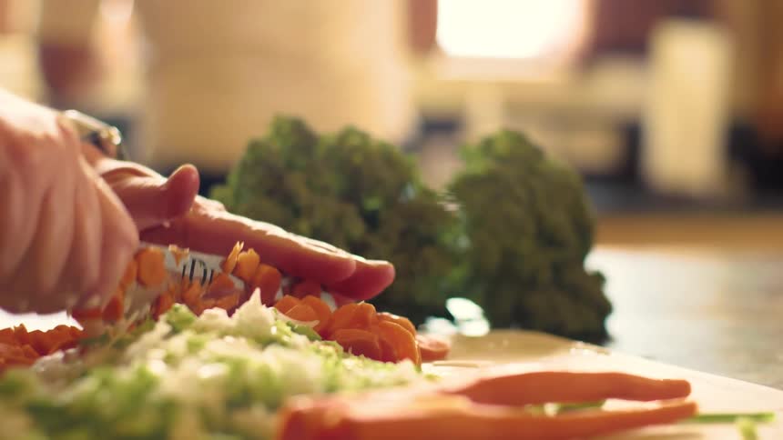 Woman using a large kitchen knife to chop and dice carrots, celery and onions on a cutting board as she prepares a meal