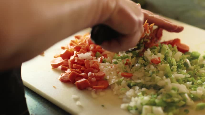Woman using a large kitchen knife to chop and dice carrots, celery and onions on a cutting board as she prepares a meal
