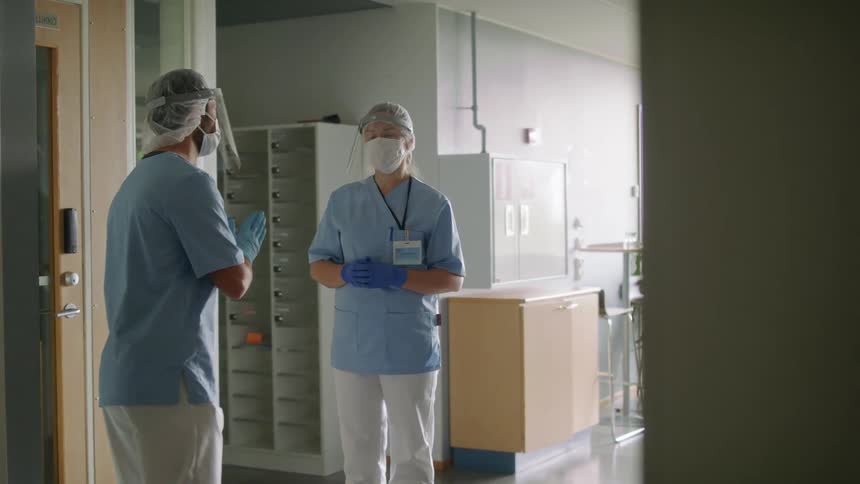 Two medical personnel in scrubs and protective gear stand in hallway talking briefly