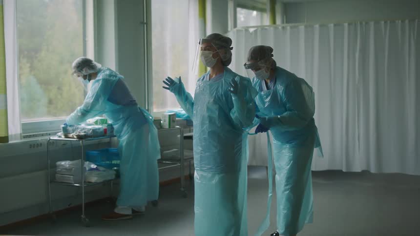 Onenurse helps another member of the surgical team tie their protective gear as a third nurse sorts through supplies