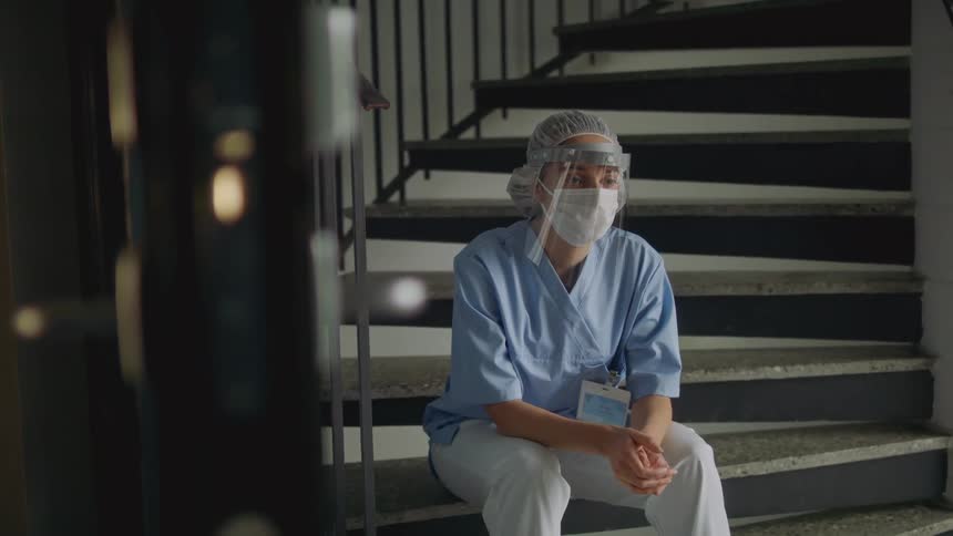 A nurse with a mask and face shield sits in a stairwell alone and nods her head