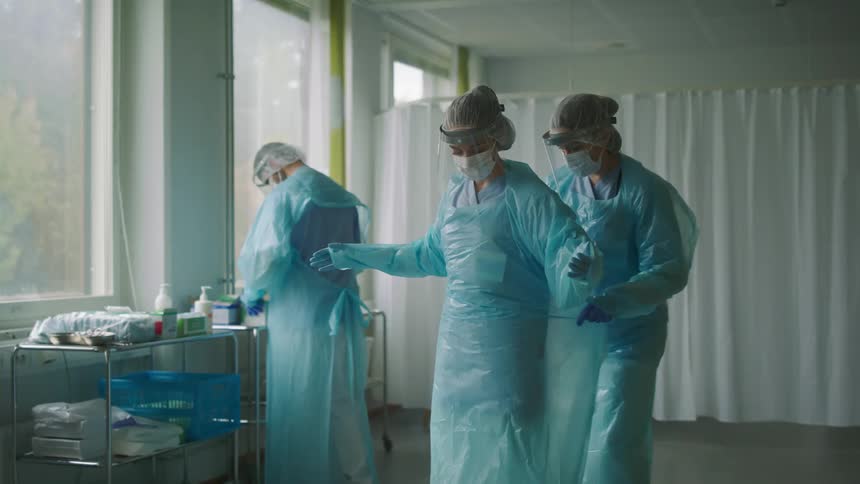 A nurse examines medical supplies on a cart while another nurse helps a woman who appears to be hyperventilating put on her medical gown