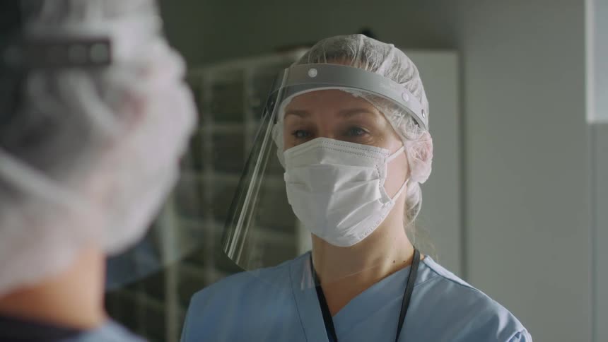 A pair of medical workers in personal protective equipment speak with one another indoors during the day