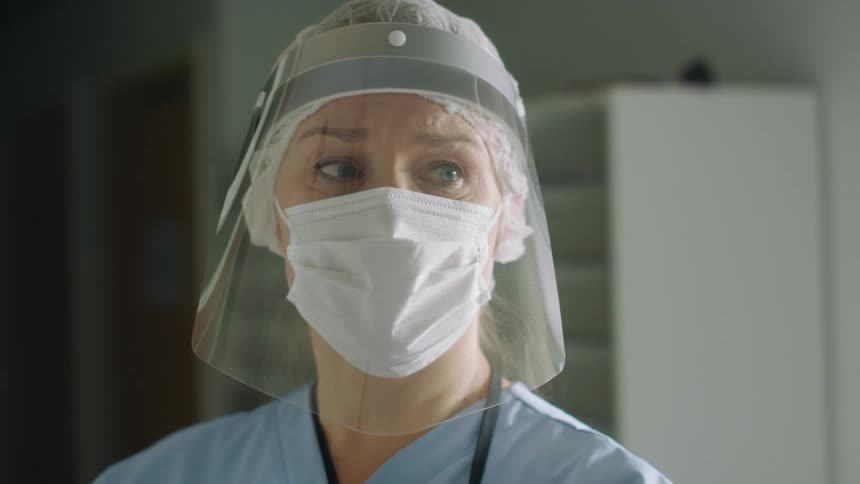 A nurse stands in a room with a mask and a face shield on