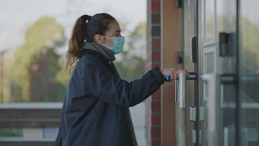 Female nurse with ponytail blue mask gray scarf and black coat opens clinic door and enters building