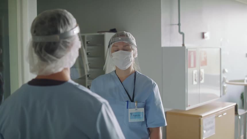 Two nurses wearing face masks and shields are standing and talking in a hospital