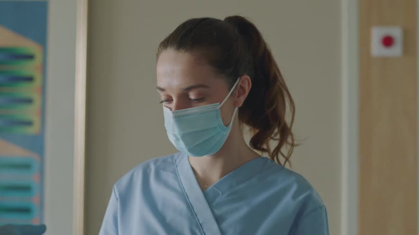 A nurse wearing scrubs and a mask stands talking in a hospital room