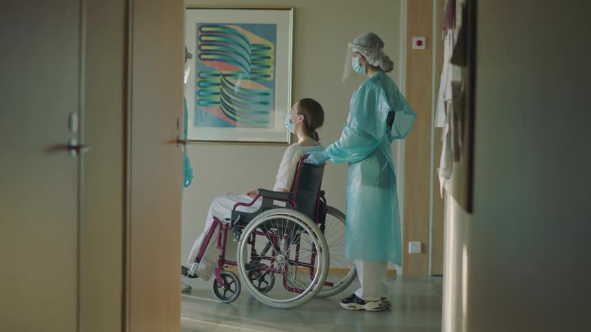 A hospital worker wearing a surgical gown and mask pushes a patient in a wheelchair out of a room and she speaks to another hospital worker