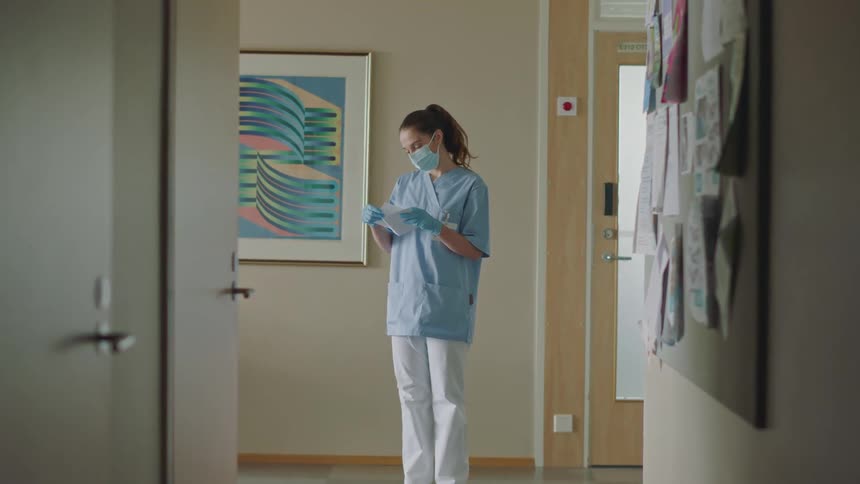 A group of doctors and nurses talks to each other and nurse with blue scrub and white pants reads a piece of paper in hospital corridor
