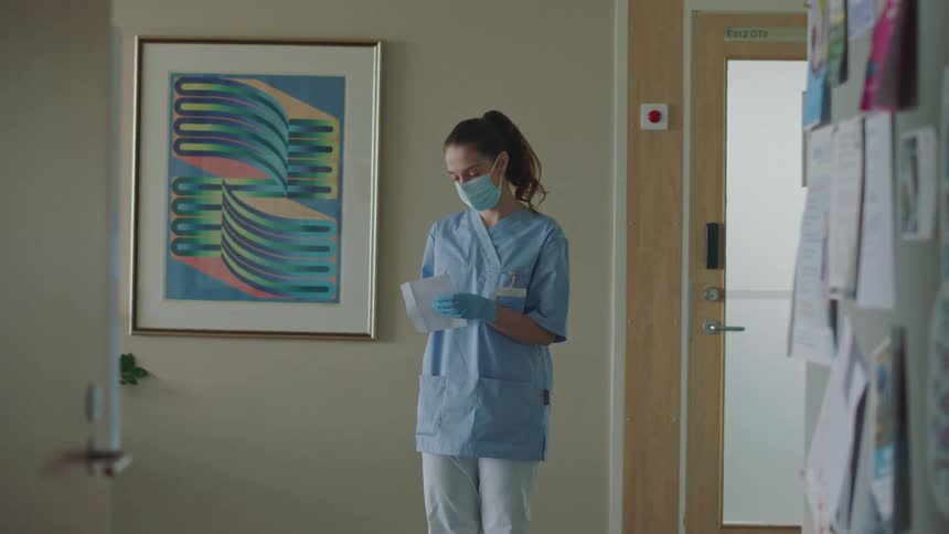 Three hospital workers wearing facemasks are standing in a room talking when another woman walks in and two of them leave as she hands an envelope to the remaining woman and she reads what is inside