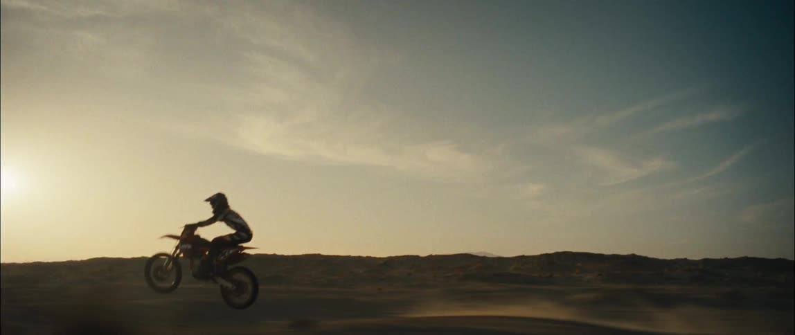 A person is riding and doing wheelies on a dirt bike on dirt hills as the sun sets