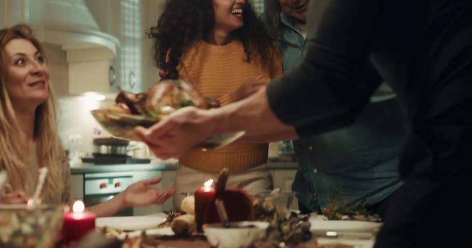 A woman carries a turkey over to the dinner table where her extended family greets her with appreciation