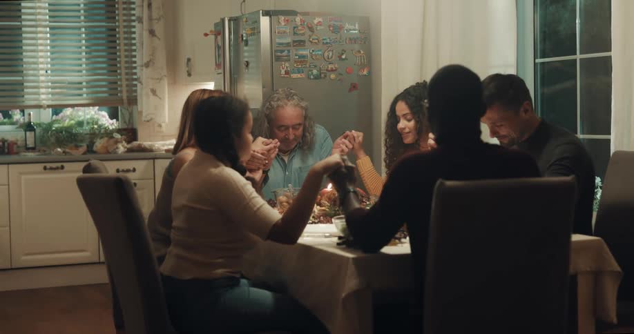 A family sitting around a dinner table holds hands while a young woman says grace