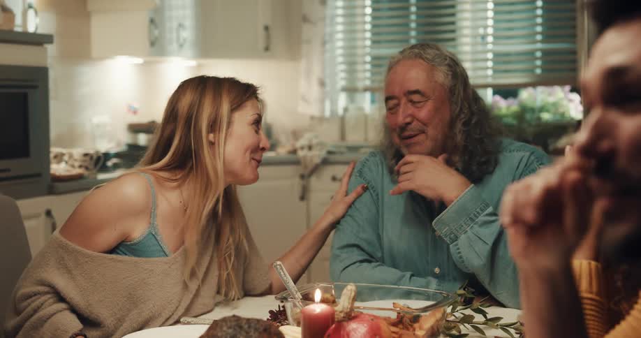 A woman leans toward the man sitting at the head of the family dinner table and caresses his arm and shoulder as they talk and smile
