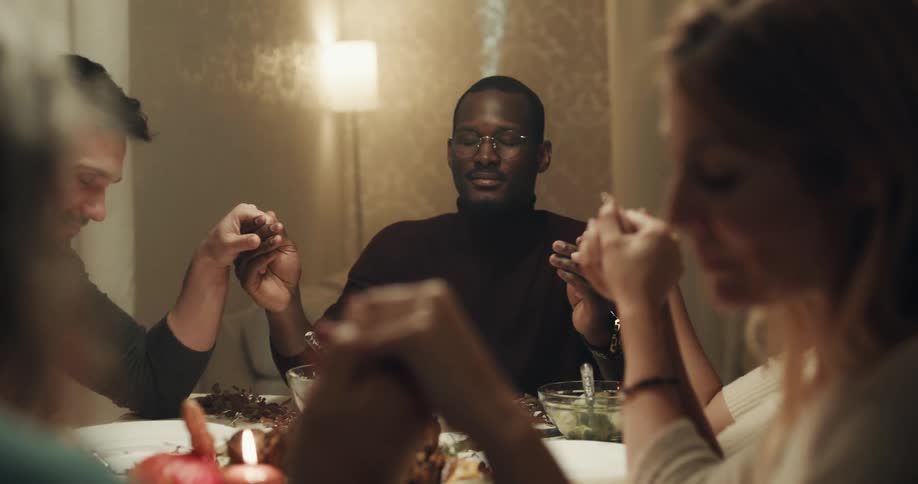 A group of people sitting around a dining table hold hands as a young woman says grace