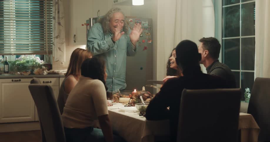 A man laughs as he stands up to carve a turkey at a dinner with friends