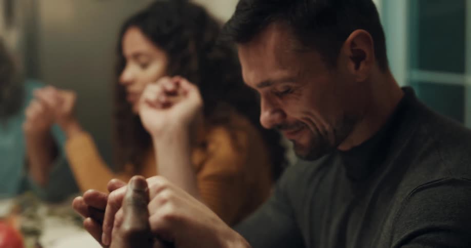 A group of friends are holding hands at a dinner table while a woman prays