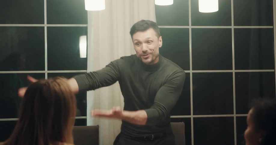 A man talking animatedly with his friends around a dining table stands to help place a roasted turkey in the center of the table