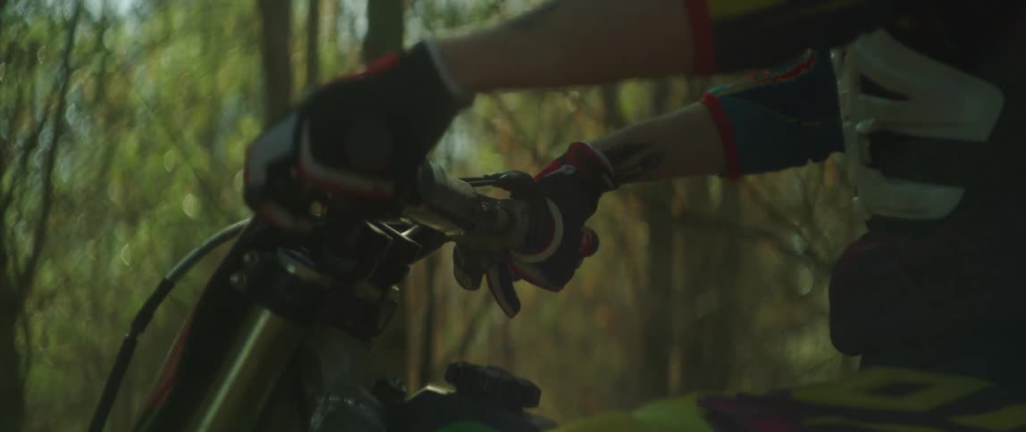 Person wearing gloves holds handlebars of dirt bike as they stand near forest