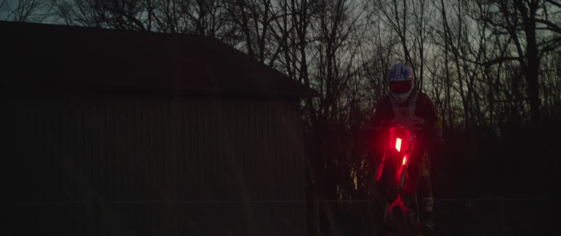 A man is riding a motorcycle with glowing red lights through a forest at dusk