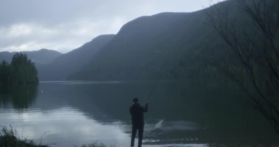 A man is fly fishing in a lake surrounded by mountains on a cloudy morning