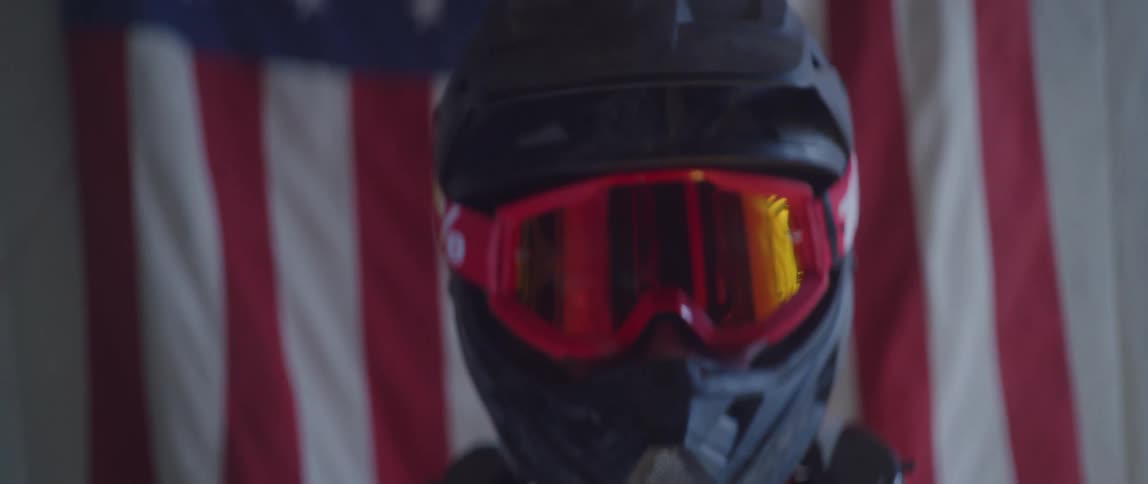 A person wearing a racing helmet and goggles is standing in front of an american flag