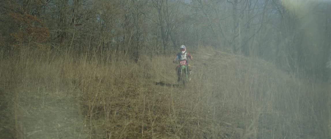A motorcyclist rides an offroad motorcycle through the grass of a woodland during the day