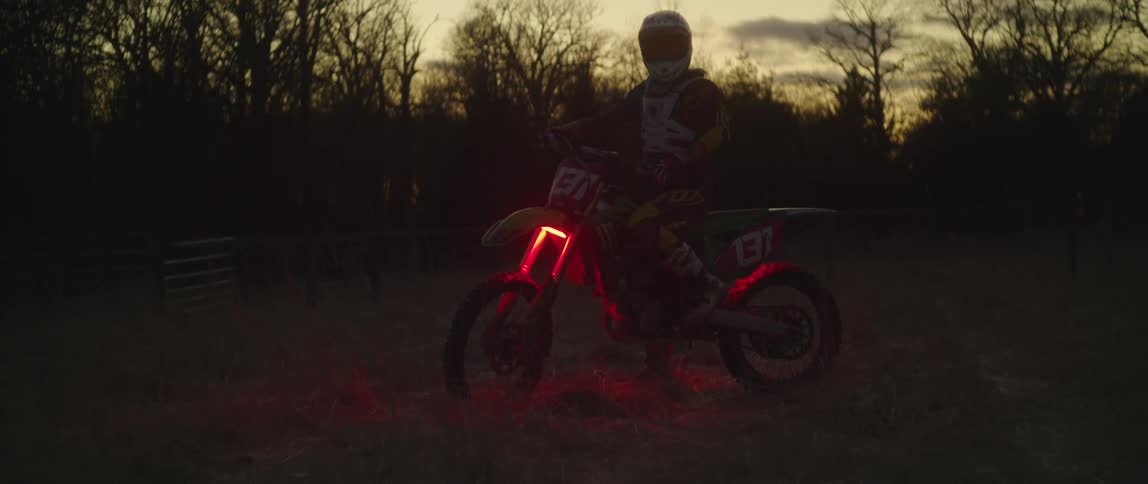 A motorcyclist looks into the camera while posing atop a dirt bike with glowing red lights in a field at sunset