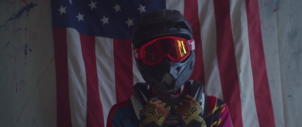 Person wearing a helmet stands in front of an american flag holding their collar with gloved hands