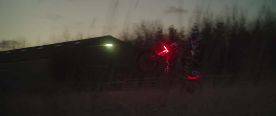 A motorcyclist popping a wheelie while riding in a rural area at dusk with red lights glowing on his dirt bike
