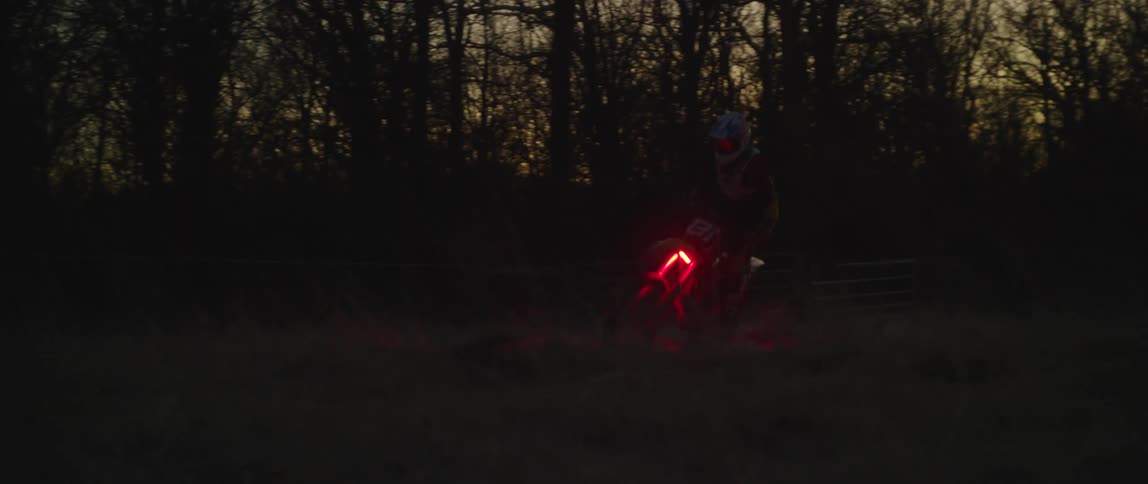 Person rides motorcycle with red lighting through field near building at night